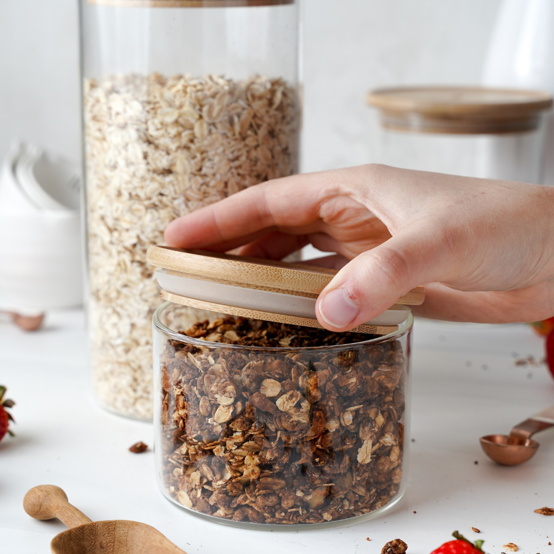 small glass jar with bamboo lid half opened containing muesli with another taller glass jar in the background